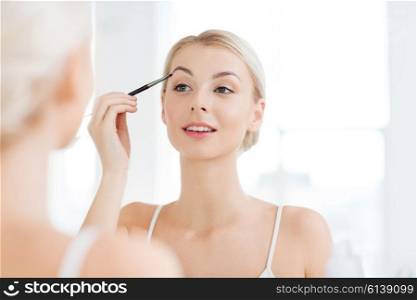 beauty, make up, cosmetics, morning and people concept - young woman applying makeup drawing eyebrow with brush and looking to mirror at home bathroom