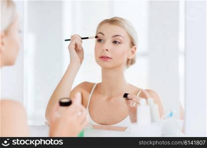 beauty, make up, cosmetics, morning and people concept - young woman applying eyeshade with makeup brush and looking to mirror at home bathroom