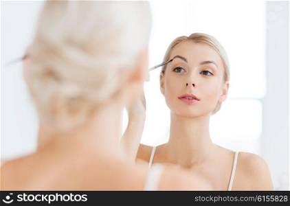 beauty, make up, cosmetics, morning and people concept - young woman applying makeup drawing eyebrow with brush and looking to mirror at home bathroom