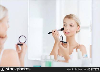 beauty, make up, cosmetics, morning and people concept - young woman applying blush with makeup brush and looking to mirror at home bathroom