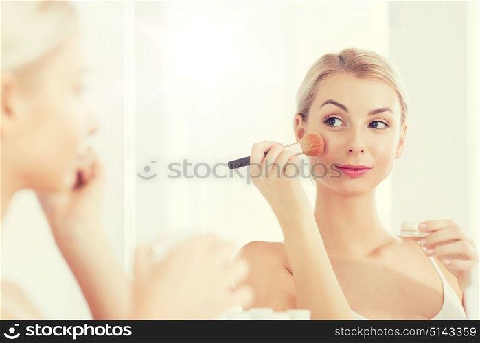 beauty, make up, cosmetics, morning and people concept - young woman makeup brush and powder foundation looking to mirror at home bathroom. woman with makeup brush and powder at bathroom