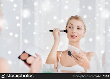 beauty, make up, cosmetics, morning and people concept - young woman with makeup brush and contouring and highlighting palette looking to mirror at home bathroom over snow