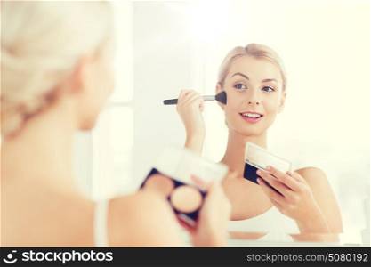 beauty, make up, cosmetics, morning and people concept - young woman with makeup brush and contouring and highlighting palette looking to mirror at home bathroom. woman with makeup brush and foundation at bathroom