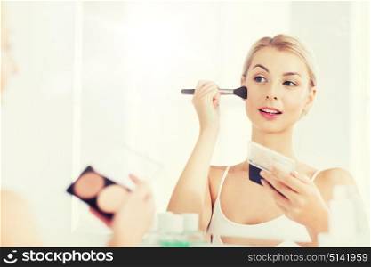 beauty, make up, cosmetics, morning and people concept - young woman with makeup brush and contouring and highlighting palette looking to mirror at home bathroom. woman with makeup brush and foundation at bathroom