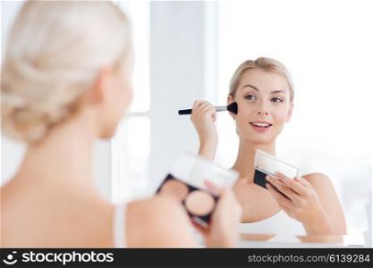 beauty, make up, cosmetics, morning and people concept - young woman with makeup brush and contouring and highlighting palette looking to mirror at home bathroom