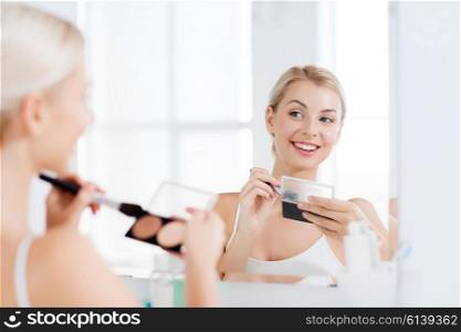 beauty, make up, cosmetics, morning and people concept - young woman with makeup brush and contouring and highlighting palette looking to mirror at home bathroom
