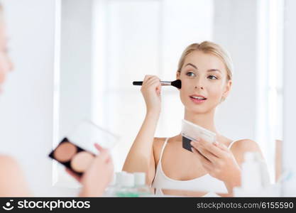 beauty, make up, cosmetics, morning and people concept - young woman with makeup brush and contouring and highlighting palette looking to mirror at home bathroom