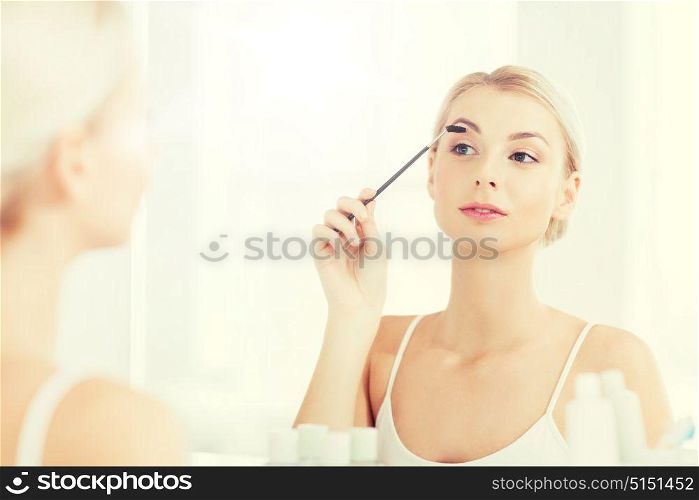 beauty, make up, morning and people concept - young woman brushing eyebrow with brush and looking to mirror at home bathroom. woman brushing eyebrow with brush at bathroom