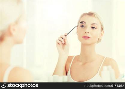 beauty, make up, morning and people concept - young woman brushing eyebrow with brush and looking to mirror at home bathroom. woman brushing eyebrow with brush at bathroom