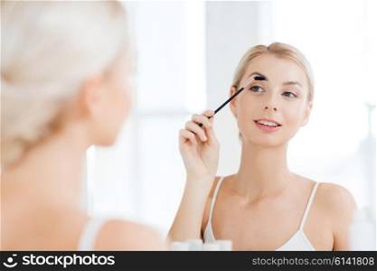 beauty, make up, morning and people concept - young woman brushing eyebrow with brush and looking to mirror at home bathroom