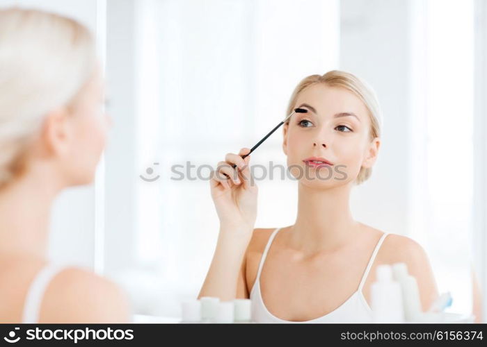 beauty, make up, morning and people concept - young woman brushing eyebrow with brush and looking to mirror at home bathroom