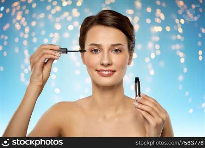 beauty, makeup and cosmetics concept - beautiful young woman applying mascara over holidays lights on blue background. beautiful woman applying mascara