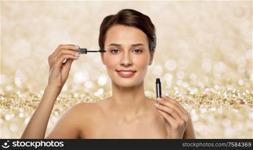 beauty, makeup and cosmetics concept - beautiful young woman applying mascara over shimmering golden glitter on background. beautiful woman applying mascara