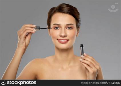 beauty, makeup and cosmetics concept - beautiful young woman applying mascara over grey background. beautiful woman applying mascara