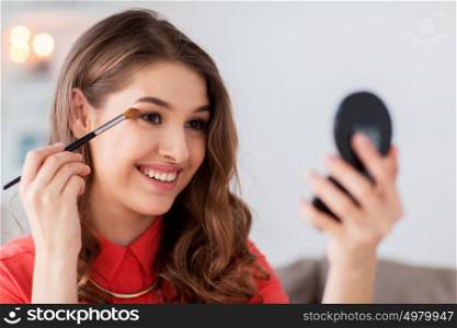 beauty, makeup, cosmetics and people concept - happy smiling young woman applying eyeshade with eye shadow brush and looking to mirror at home. woman with eye shadow brush and mirror does makeup