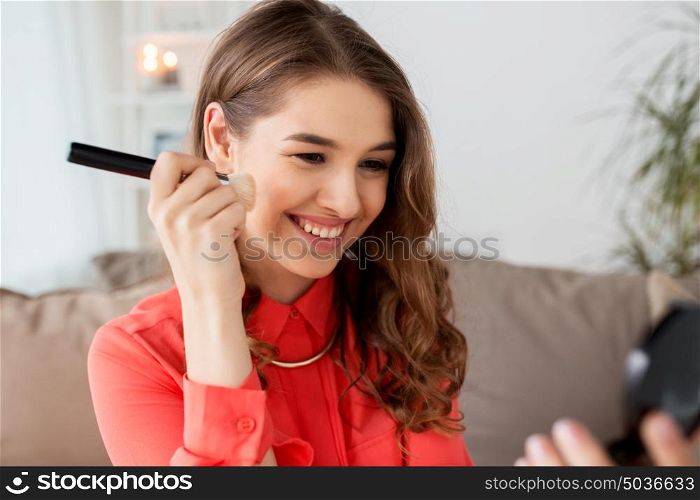 beauty, makeup, cosmetics and people concept - happy smiling young woman with brush applying bronzer or blush to her face and looking to mirror at home. woman with eye shadow brush and mirror does makeup