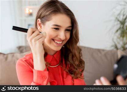 beauty, makeup, cosmetics and people concept - happy smiling young woman with brush applying bronzer or blush to her face and looking to mirror at home. woman with eye shadow brush and mirror does makeup