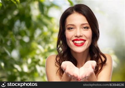 beauty, organic, make up and people concept - happy smiling young woman with red lipstick holding something imaginary on palms over green natural background. beautiful smiling young woman with red lipstick