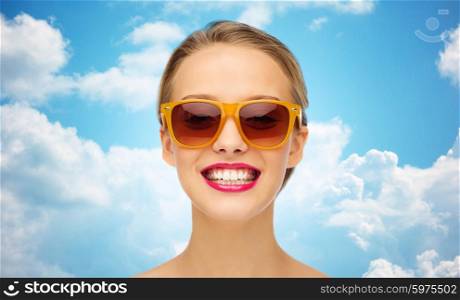 beauty, people, accessory and fashion concept - smiling young woman in sunglasses with pink lipstick on lips over blue sky and clouds background