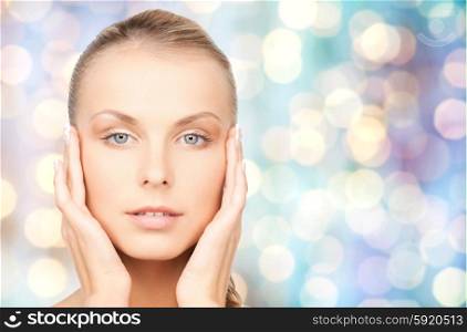 beauty, people and body care concept - beautiful young woman touching her face over blue holidays lights background