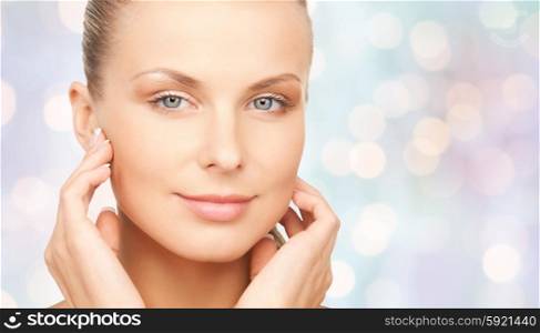beauty, people and body care concept - beautiful young woman touching her face over blue holidays lights background