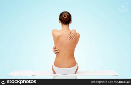 beauty, people and bodycare concept - beautiful topless young woman in white panties sitting on towel from back over blue background. beautiful topless young woman on towel from back