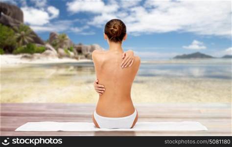 beauty, people and bodycare concept - beautiful topless young woman in white panties sitting on towel from back over exotic beach background. beautiful woman with bare top on beach