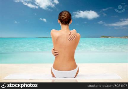 beauty, people and bodycare concept - beautiful topless young woman in white panties sitting on towel from back over exotic tropical beach background. beautiful topless woman with towel on summer beach