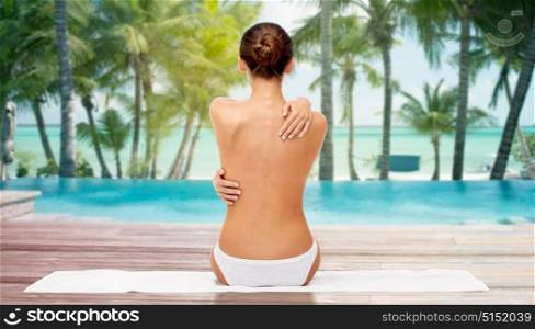 beauty, people and bodycare concept - beautiful topless young woman in white panties sitting on towel from back over beach and outdoor swimming pool background. beautiful woman with bare top on beach