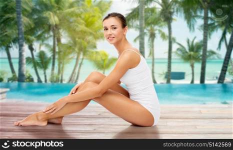 beauty, people and bodycare concept - beautiful woman touching her smooth bare legs over beach and outdoor swimming pool background. beautiful woman touching her bare legs on beach