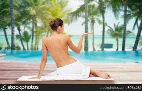 beauty, people and bodycare concept - beautiful young woman in white towel with bare top holding something invisible over beach and outdoor swimming pool background. beautiful woman in towel with bare top on beach. beautiful woman in towel with bare top on beach