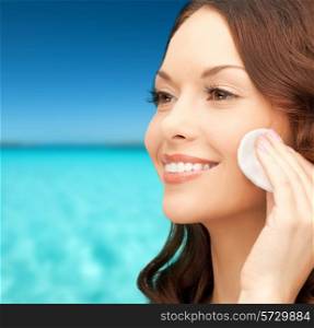 beauty, people and health concept - beautiful smiling woman cleaning face skin with cotton pad over blue sea and sky background