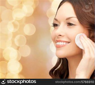 beauty, people and health concept - beautiful smiling woman cleaning face skin with cotton pad over beige lights background