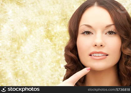 beauty, people and health concept - beautiful young woman pointing finger to her chin over yellow lights background