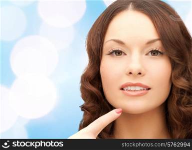 beauty, people and health concept - beautiful young woman pointing finger to her chin over blue lights background