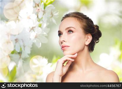 beauty, people and health concept - beautiful young woman touching her face over natural spring cherry blossom background