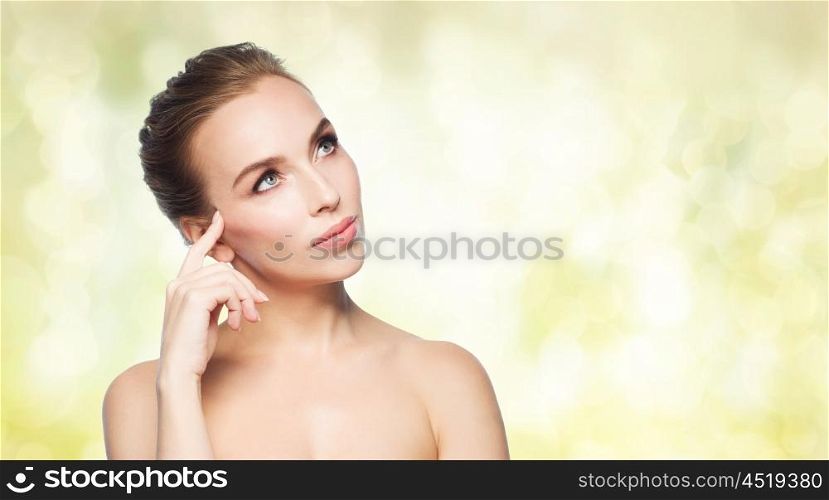 beauty, people and health concept - beautiful young woman touching her face over yellow holidays lights background