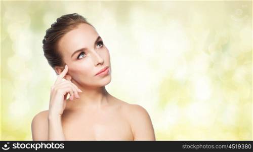 beauty, people and health concept - beautiful young woman touching her face over yellow holidays lights background