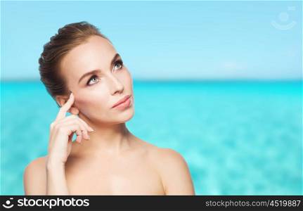 beauty, people and health concept - beautiful young woman touching her face over blue sea and sky background