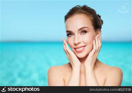 beauty, people and health concept - beautiful young woman touching her face over blue sea and sky background