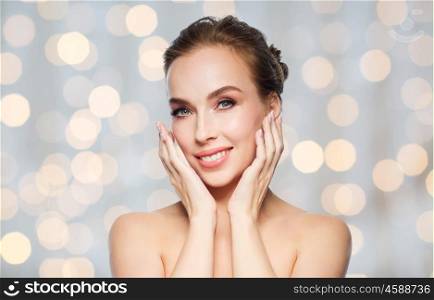 beauty, people and health concept - beautiful young woman touching her face over holidays lights background