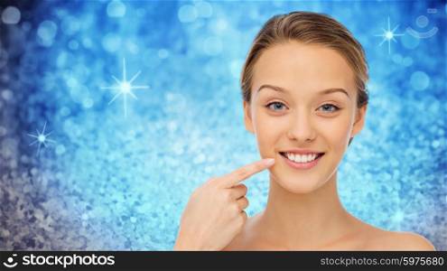 beauty, people and health concept - happy young woman pointing finger to her smile or teeth over blue holidays lights or glitter background