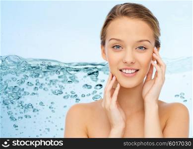 beauty, people and health concept - smiling young woman with bare shoulders touching her face over water splash on blue background