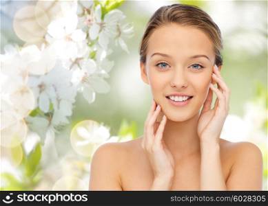 beauty, people and health concept - smiling young woman with bare shoulders touching her face over green natural background with cherry blossom