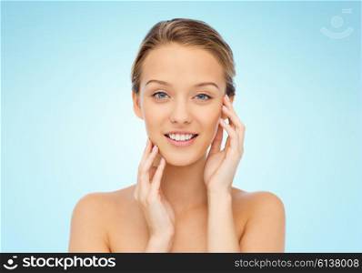 beauty, people and health concept - smiling young woman with bare shoulders touching her face over blue background