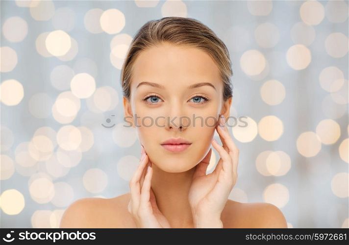 beauty, people and health concept - young woman with bare shoulders touching her face over holidays lights background