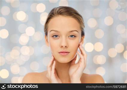 beauty, people and health concept - young woman with bare shoulders touching her face over holidays lights background