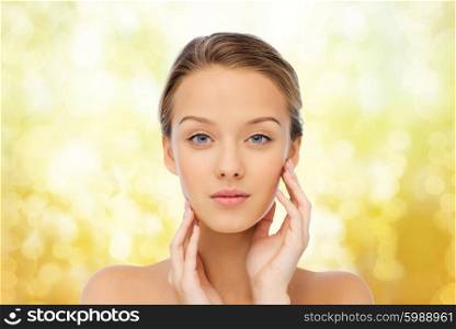 beauty, people and health concept - young woman with bare shoulders touching her face over yellow holidays lights background