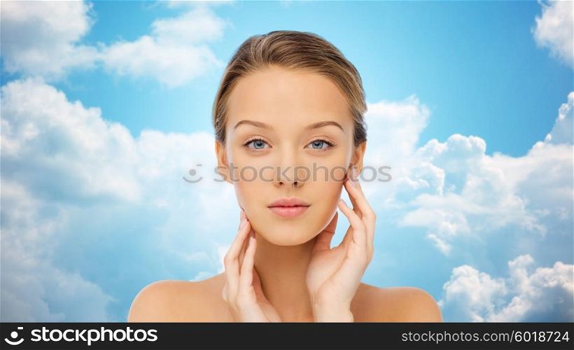 beauty, people and health concept - young woman with bare shoulders touching her face over blue sky and clouds background