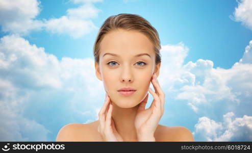 beauty, people and health concept - young woman with bare shoulders touching her face over blue sky and clouds background